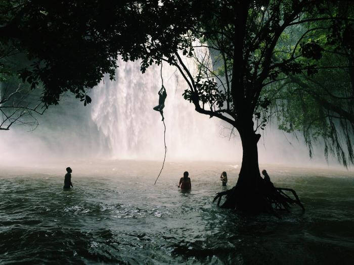 Colored photograph of a shadowy scene from the trees of people enjoying a waterfall and a swinging rope.
